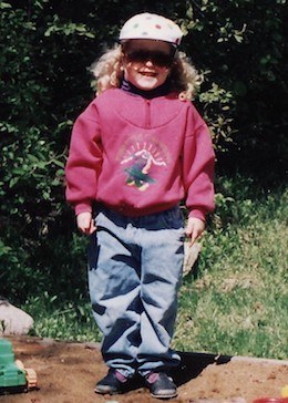 A young Alyssa wearing jeans and a pink sweatshirt, standing in a sandbox